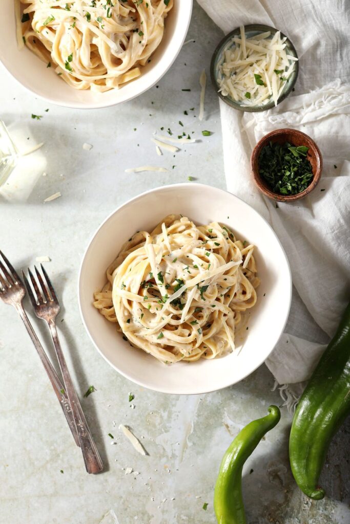 A bowl of creamy pasta topped with chopped parsley and shredded parmesan next to peppers