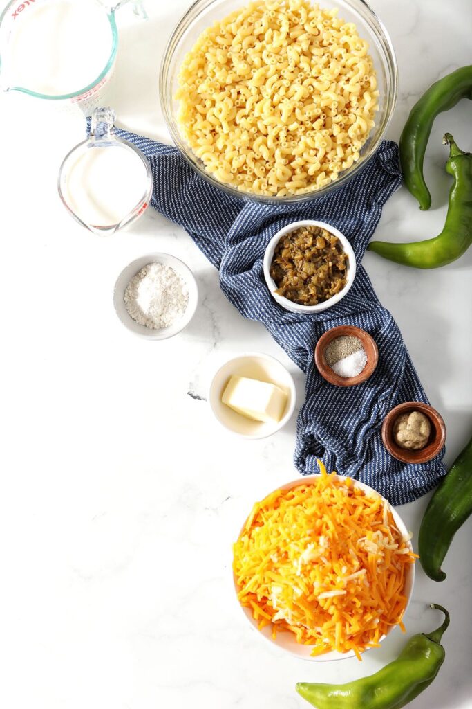 Ingredients to make macaroni and cheese in bowls on marble surrounded by green Chile peppers