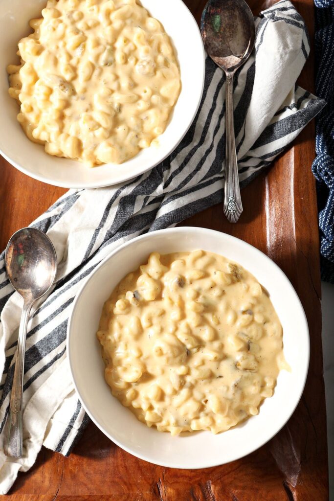 Two bowls of Hatch Chile macaroni and cheese sit on a wooden platter with spoons and a striped towel