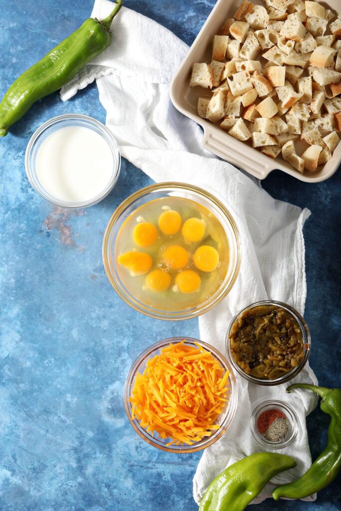 Ingredients to make a Hatch Chile egg casserole sit on a blue countertop in bowls with fresh peppers