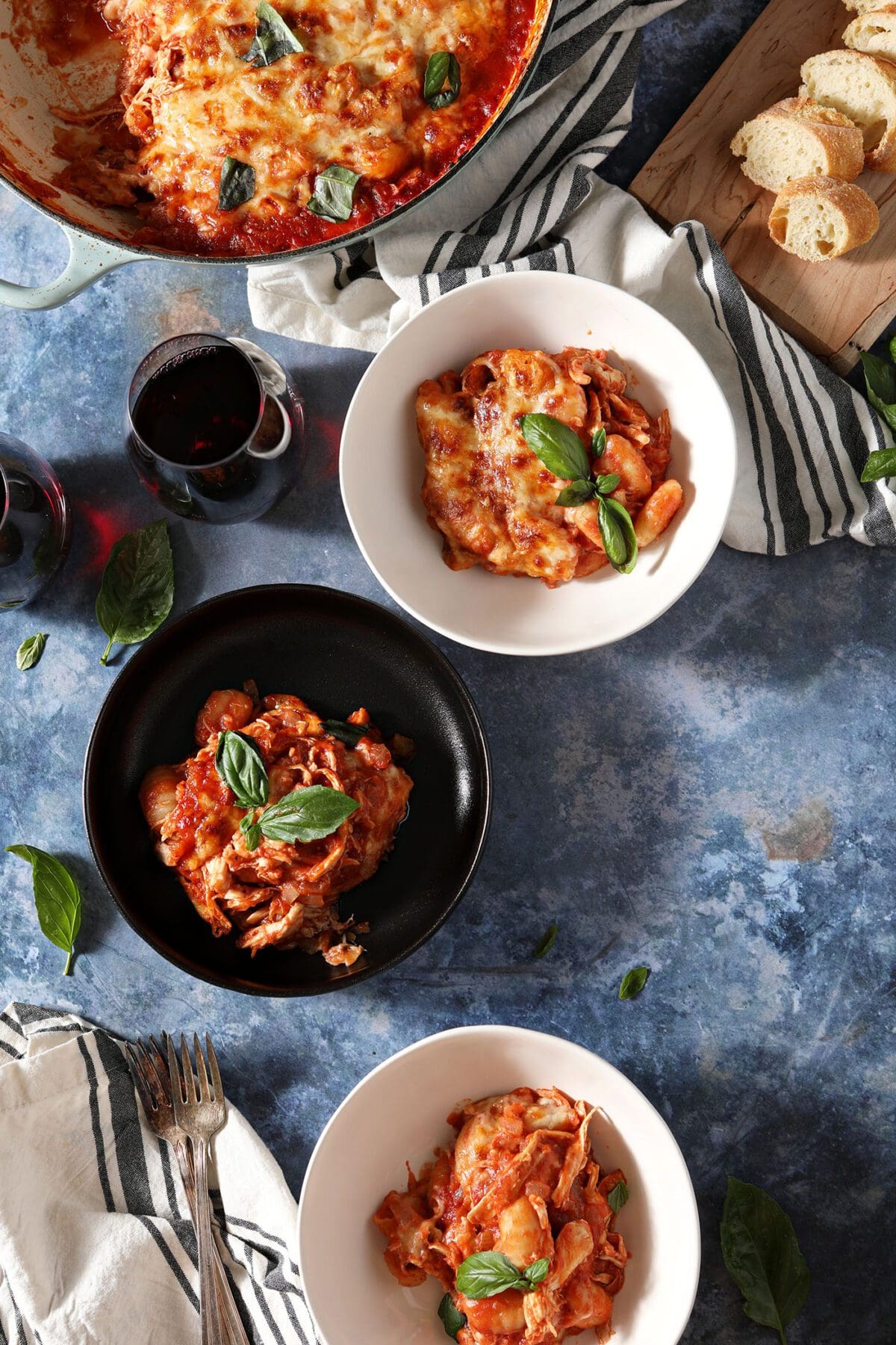 Three bowls of a gnocchi bake with chicken on a blue table with forks, wine, bread and the baking dish