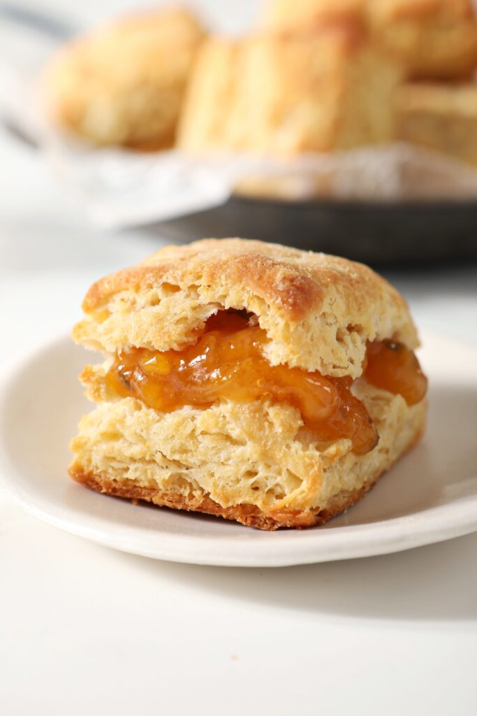 A butter biscuit filled with peach jam on a white plate in front of a bowl of biscuits