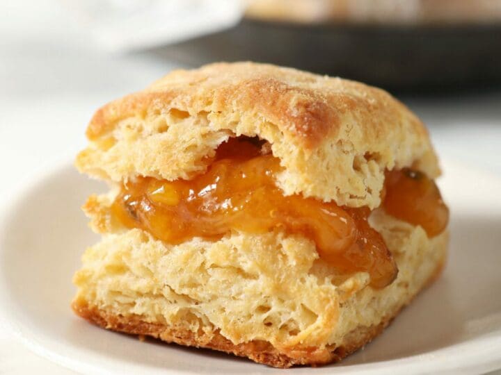 A butter biscuit filled with peach jam on a white plate in front of a bowl of biscuits