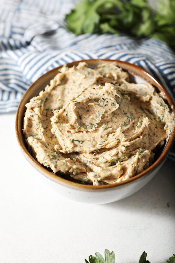 Cajun Butter in a white bowl with a brown rim on marble next to a blue hatched towel and herbs