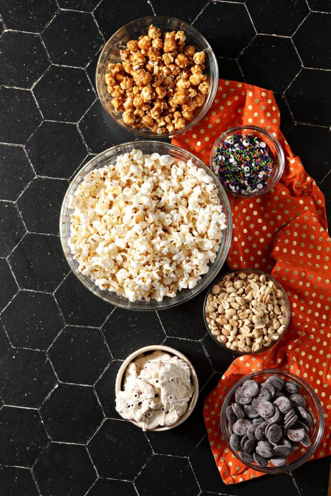 Bowls holding ingredients to make a sweet and salty Halloween popcorn snack mix on a black countertop