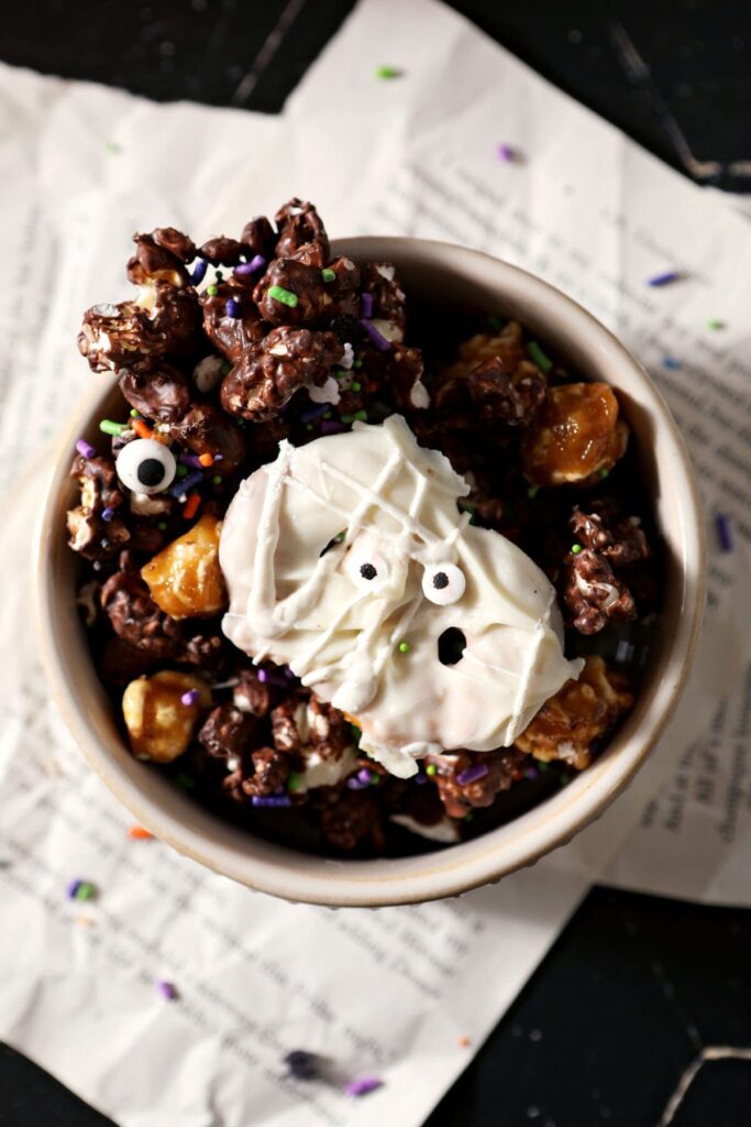 Close up of a white bowl holding Halloween Popcorn Snack mix on top of papers