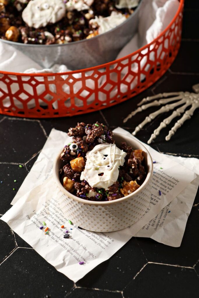 A small bowl of Halloween popcorn snack mix with chocolate and caramel and white chocolate pretzels next to a larger bowl