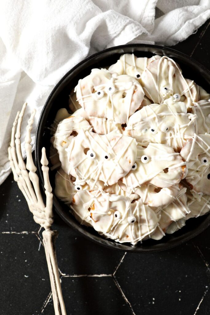 Halloween pretzels decorated like mummies in a black bowl next to a plastic skeleton hand