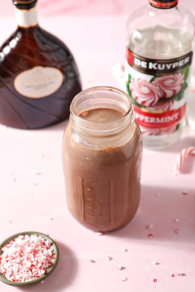 A jar of hot cocoa with bottles of peppermint schnapps and white chocolate liqueur on a pink surface