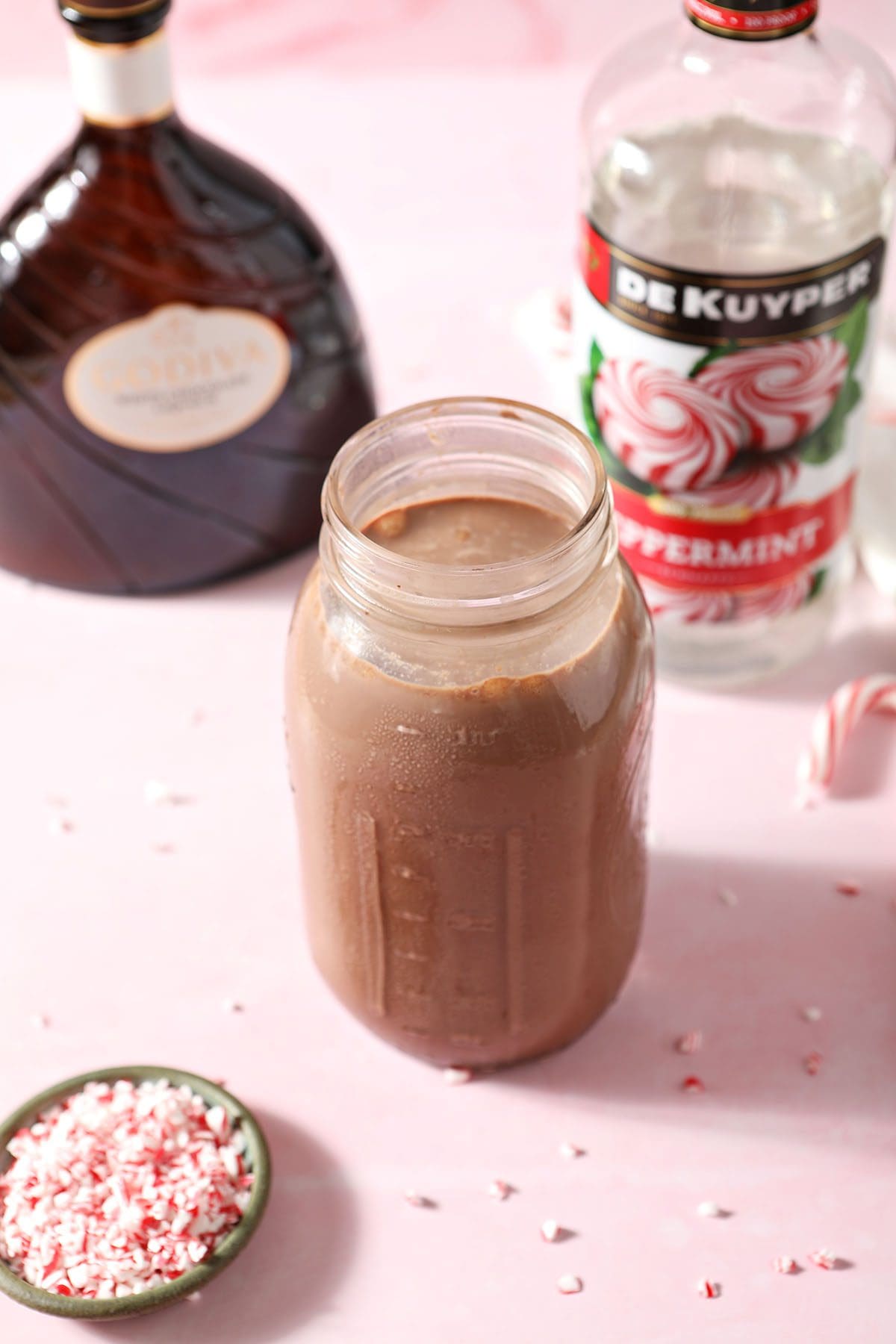 A jar of hot cocoa with bottles of peppermint schnapps and white chocolate liqueur on a pink surface