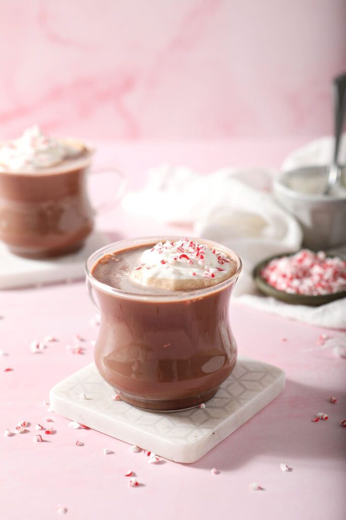 Two mugs of Peppermint Patty drinks on marble coasters on a pink surface garnished with sweetened whipped cream and crushed candy cane