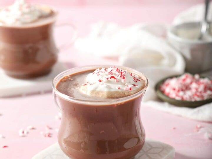 Two mugs of Peppermint Patty drinks on marble coasters on a pink surface garnished with sweetened whipped cream and crushed candy cane