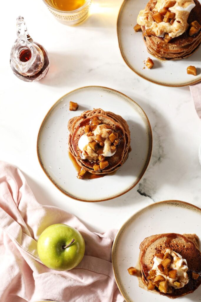 Three plates of apple pancakes from above garnished with caramelized apples, syrup and whipped cream