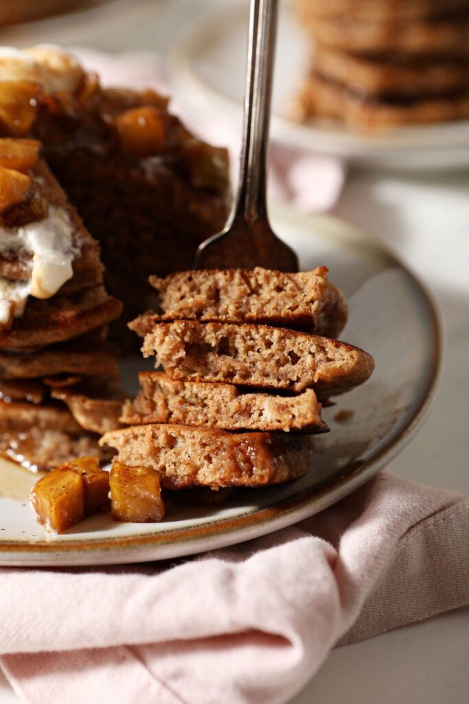 A fork holds a bite of Apple Cider Pancakes next to a stack of pancakes