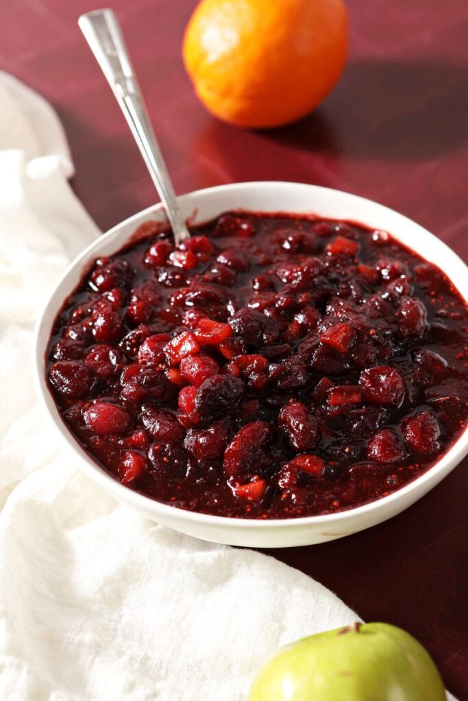 A white bowl of cranberry chutney with a spoon in it on a red countertop with a white towel