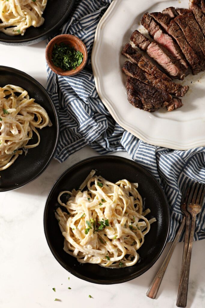 Bowls of fettuccine pasta in Alfredo next to a plate of sliced steak