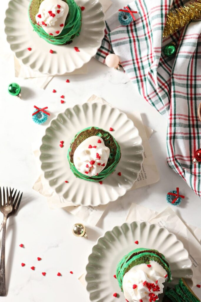 Three plates of festive Christmas pancakes on white plates surrounded by cheerful Christmas decorations and heart sprinkles