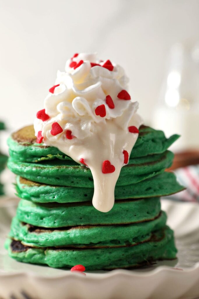 Close up of green pancakes topped with whipped cream and cream cheese frosting with red heart sprinkles