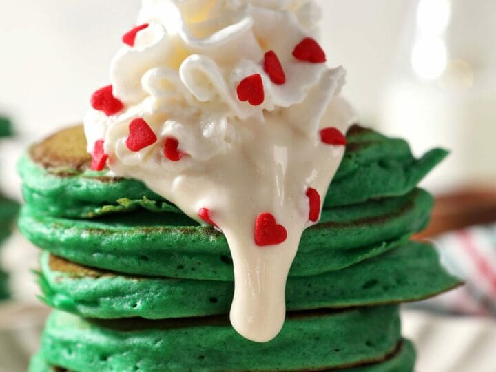 Close up of green pancakes topped with whipped cream and cream cheese frosting with red heart sprinkles