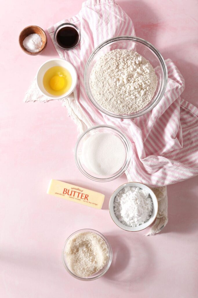 Ingredients to make no chill drop sugar cookies in bowls on a pink countertop with a pink striped towel