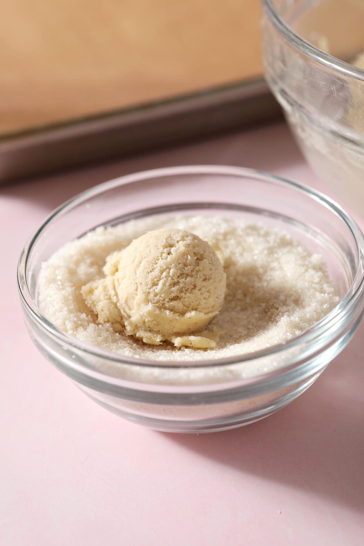 A ball of sugar cookie dough in a bowl of vanilla sugar by a parchment-lined sheet pan