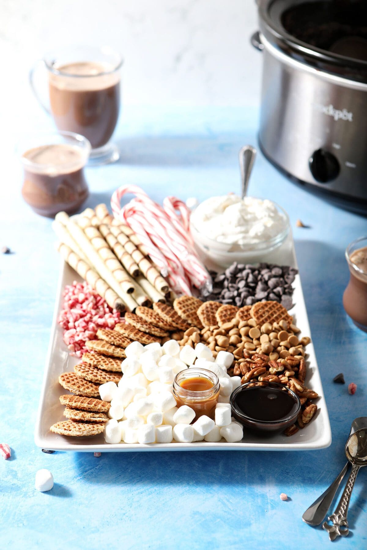 A hot cocoa charcuterie board on a blue countertop next to a slow cooker full of cocoa