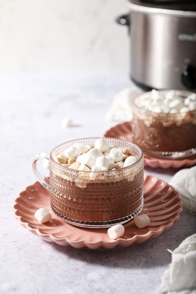 Two clear mugs of Slow Cooker Hot Cocoa topped with mini marshmallows on pink plates