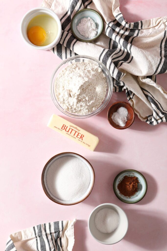 Ingredients to make cookies on a pink countertop with a gray and white striped towel