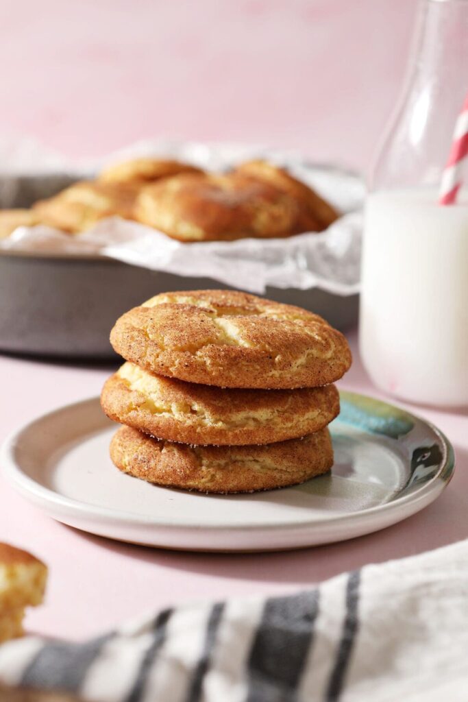 Soft and fluffy Snickerdoodle Cookies are full of comfort and deliciousness. Covered in cinnamon sugar, no one can resist these sweet, flavorful cookies.  Three cookies stacked on a plate next to a carton of milk