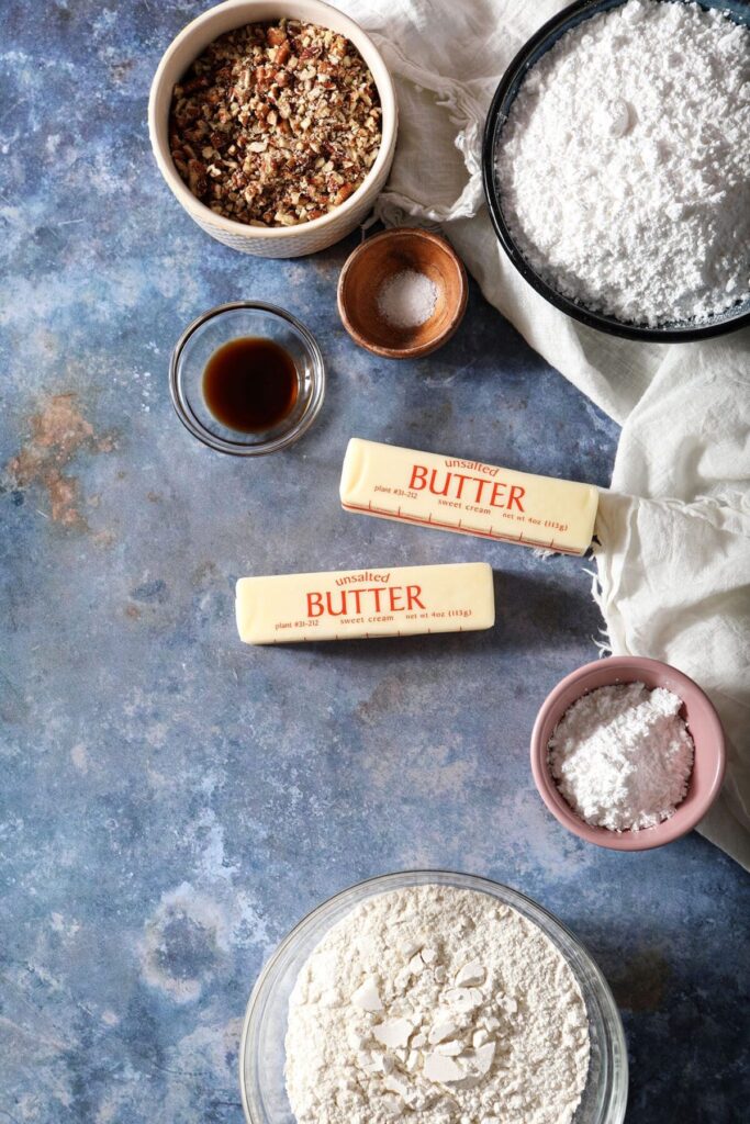 Ingredients to make snowball cookies on a blue countertop with a white tea towel