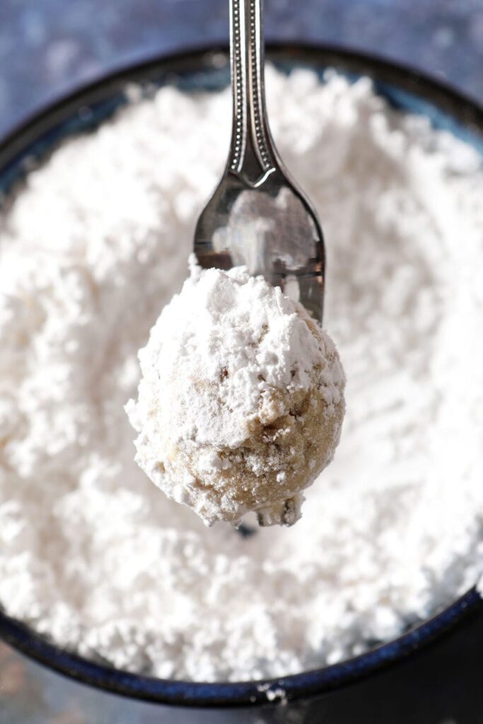 A fork holds a snowball cookie over a bowl of powdered sugar