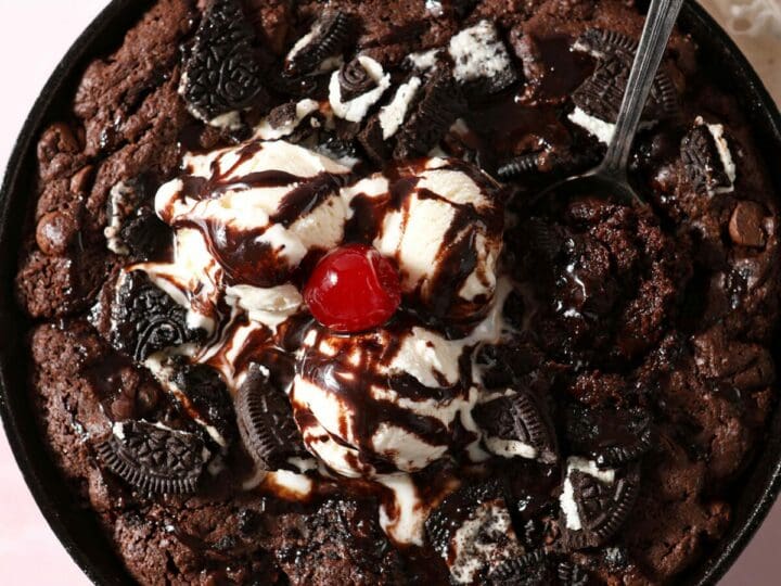 A spoon scoops into a Oreo skillet brownie topped with ice cream, chocolate sauce and a cherry
