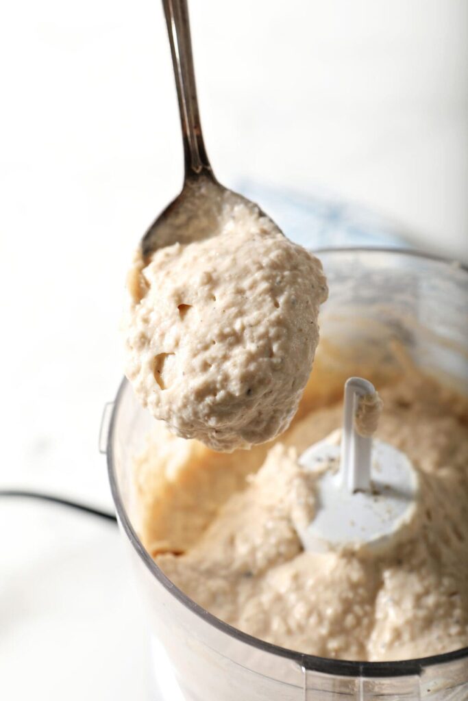 A spoon holds a scoop of Roasted Garlic Hummus above a food processor