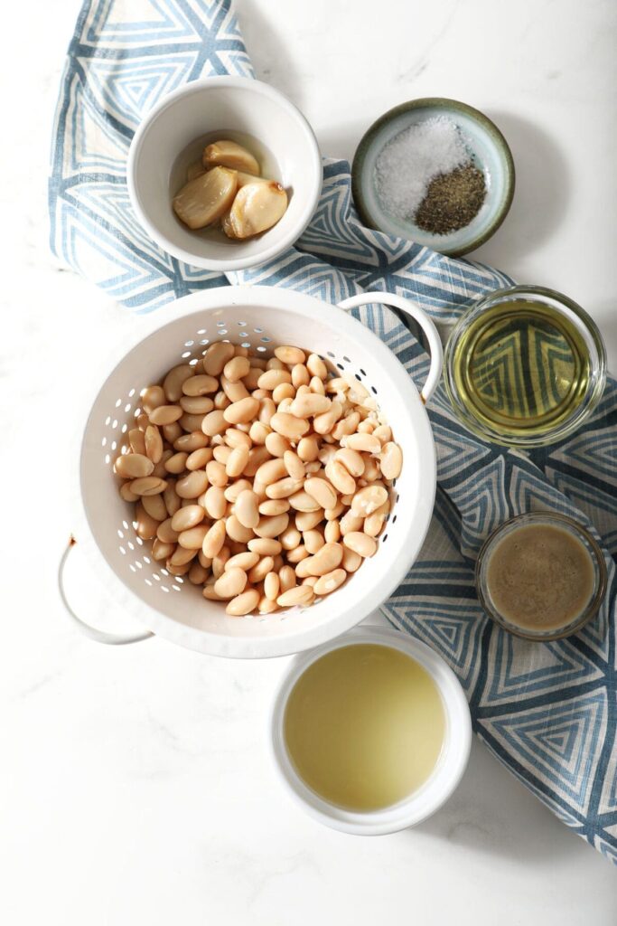 Ingredients to make Roasted Garlic Hummus with white beans in bowls on a marble countertop with a geometric blue towel