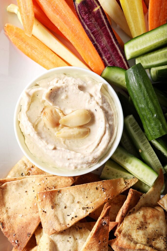 A serving bowl of Roasted Garlic White Bean Hummus surrounded by homemade pita chips, cucumbers and carrots