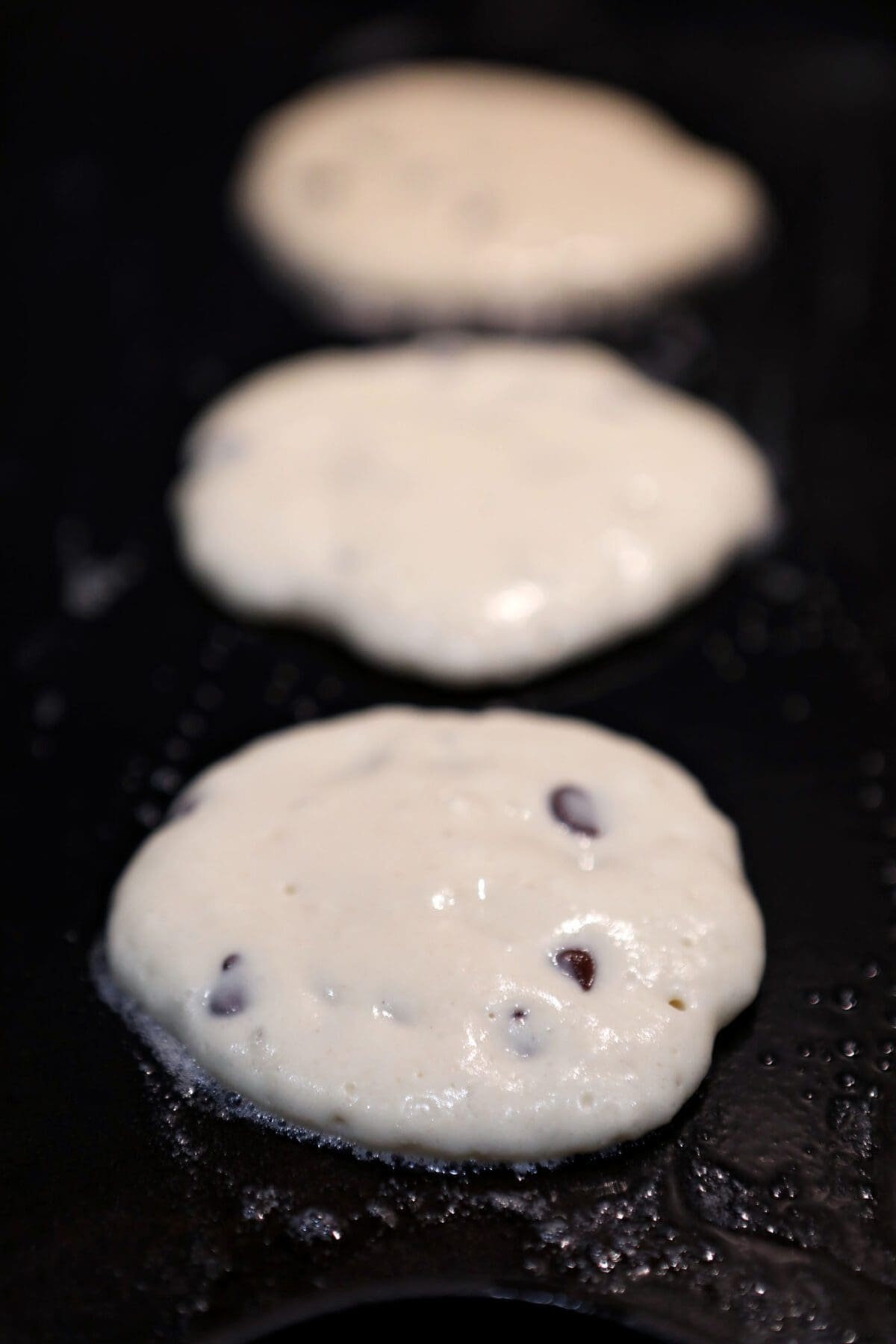 Three homemade pancakes with chocolate chips on a griddle cooking