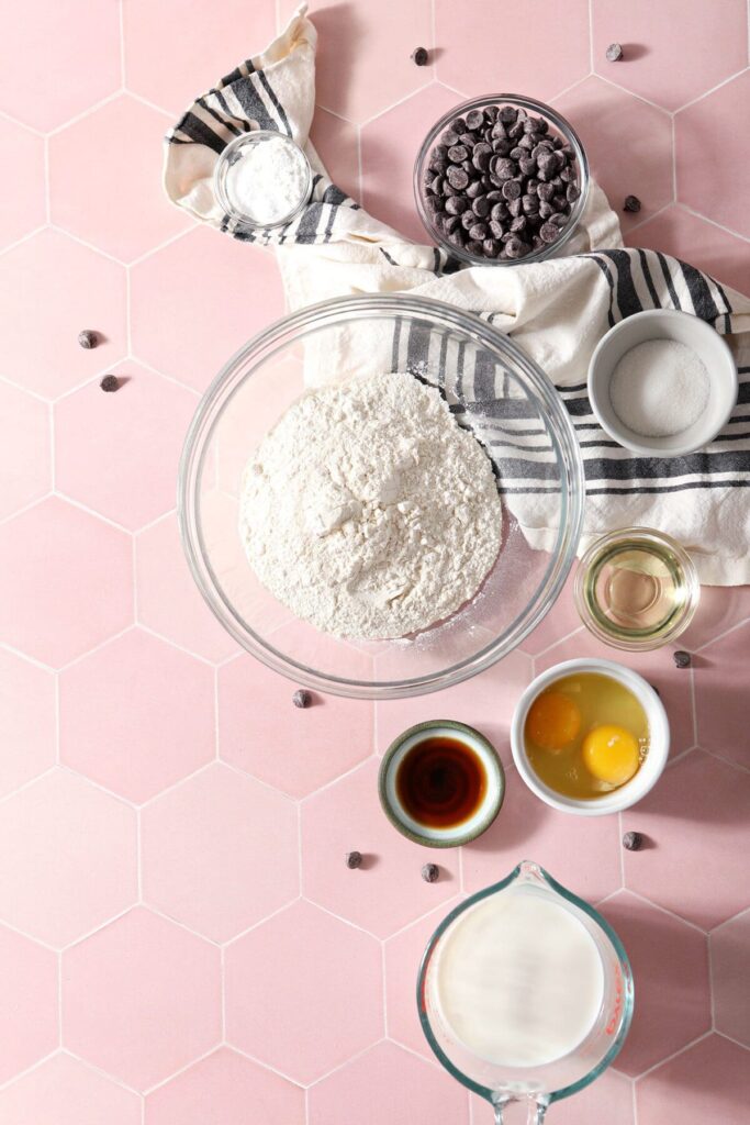 Ingredients for homemade pancakes with chocolate chips in bowls on a pink tile countertop