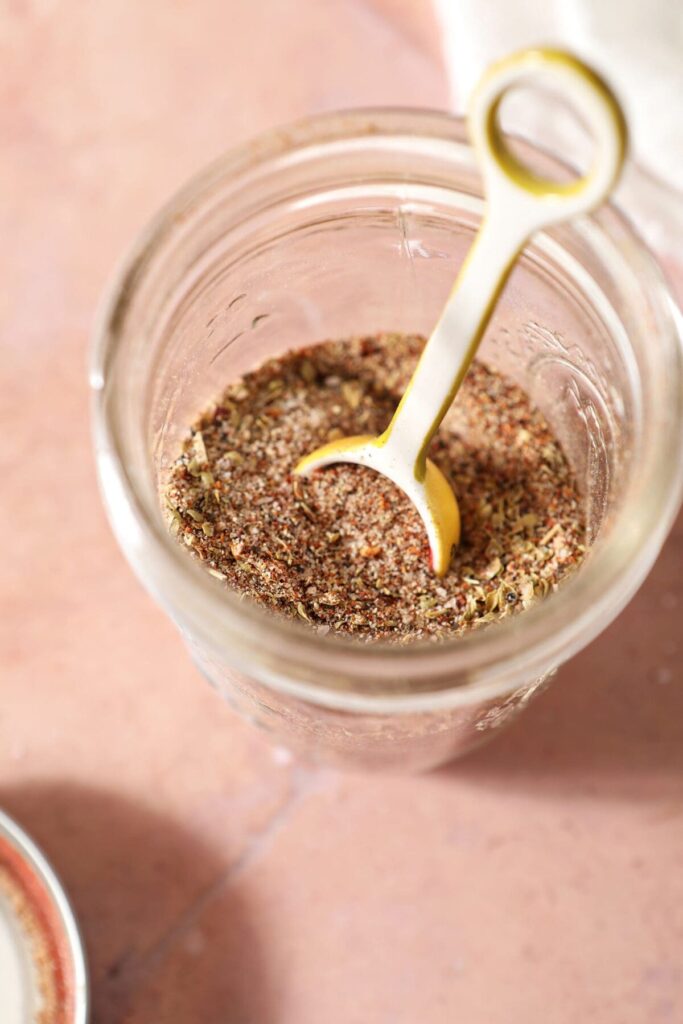 Creole seasoning in a jar with a teaspoon in the jar on a red tile