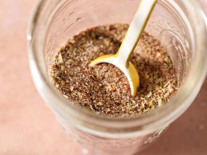 Creole seasoning in a jar with a teaspoon in the jar on a red tile