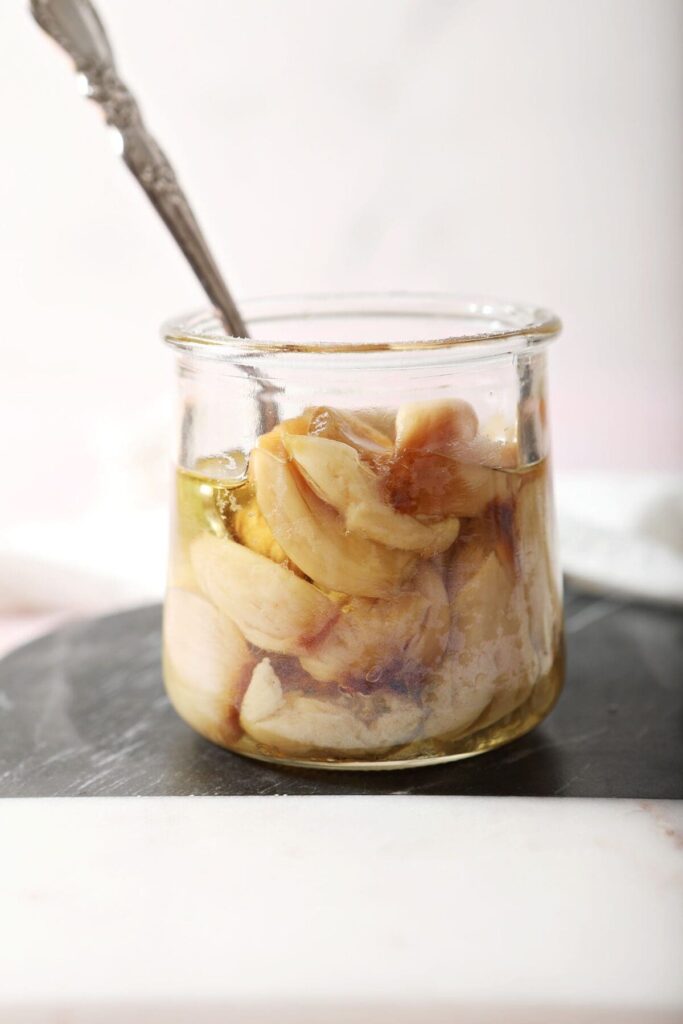 A jar of roasted garlic cloves with a spoon inside sits on black and white cutting board