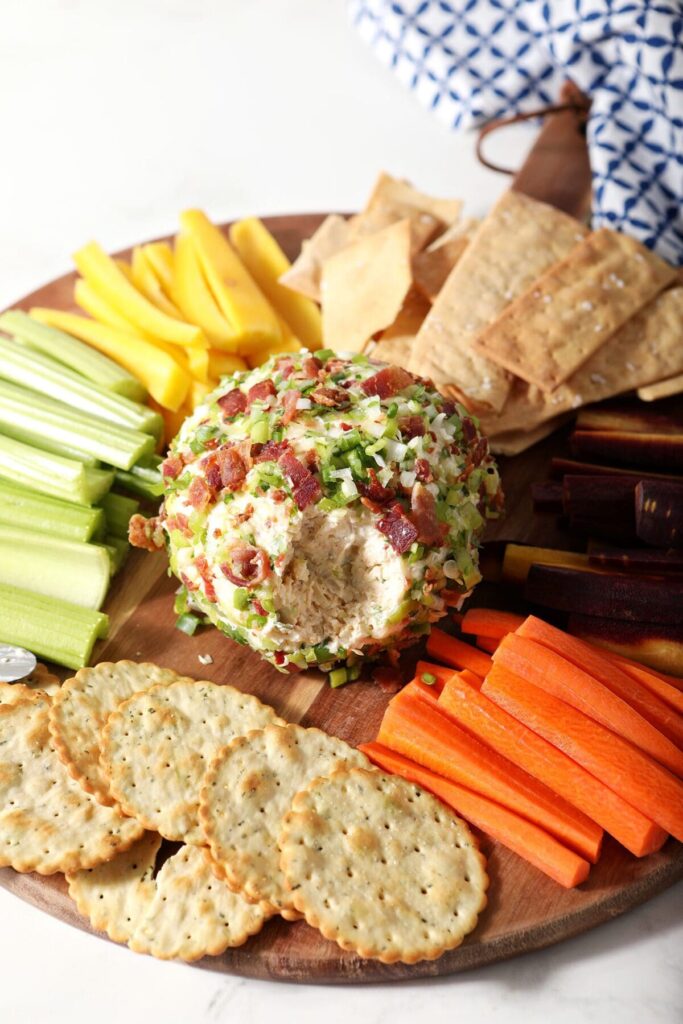 A cheeseball coated in green onions and bacon in the middle of a wooden board with crackers and raw vegetables