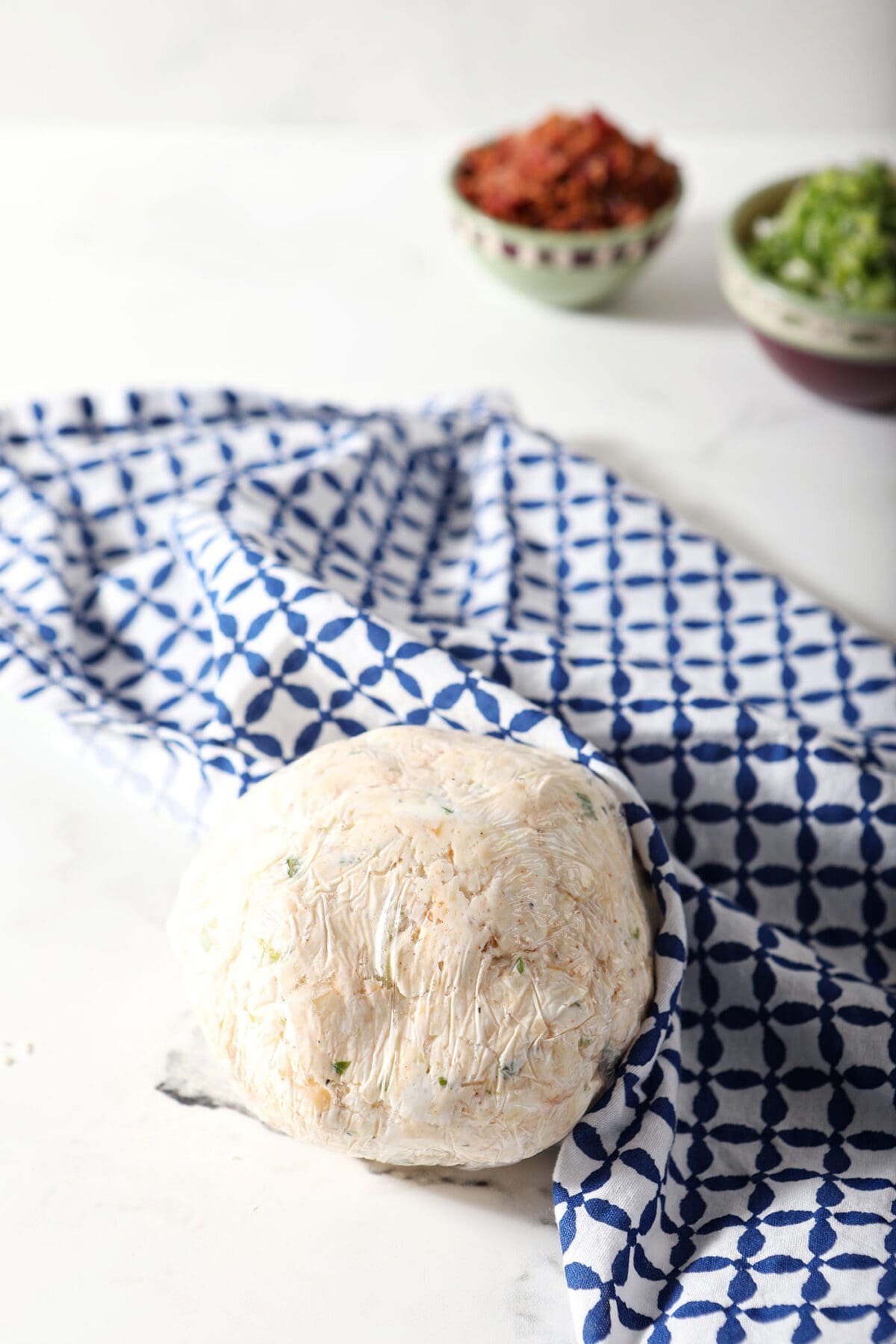 A cheeseball wrapped in plastic wrap with a blue patterned towel on marble