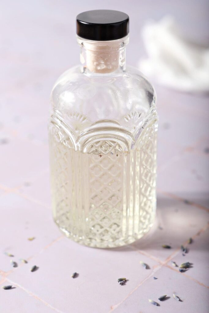 A bottle of lavender syrup on a purple tiled backdrop surrounded by dried lavender