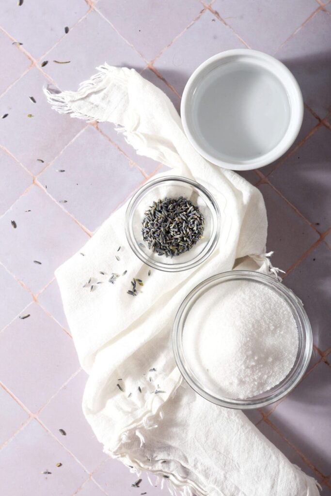 Ingredients to make lavender simple syrup in bowls on a white towel on a purple tiled backdrop