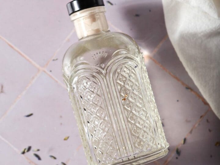 A bottle of lavender syrup on a purple lies on purple tile with dried lavender and a cream-colored linen