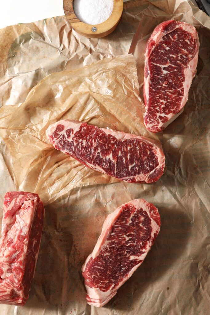 Four raw steaks on brown butcher’s paper next to a wooden container of salt