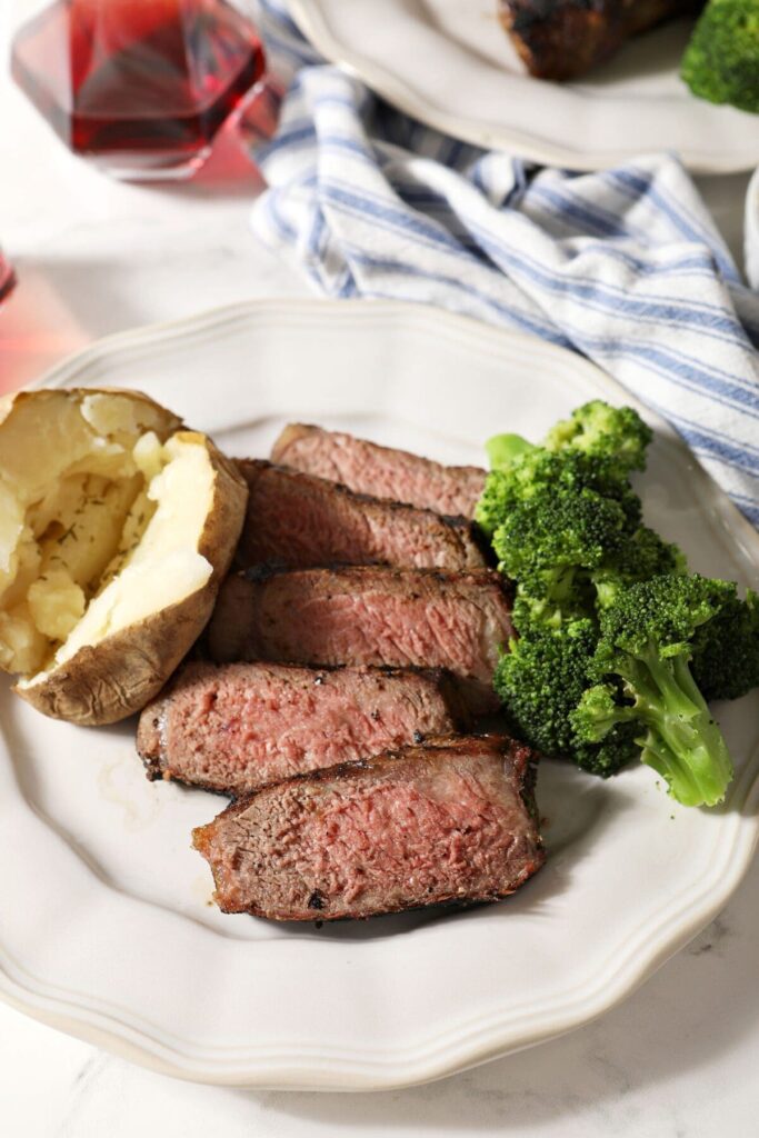 Close up of a white plate of sliced grilled steak with steamed broccoli and a baked potato