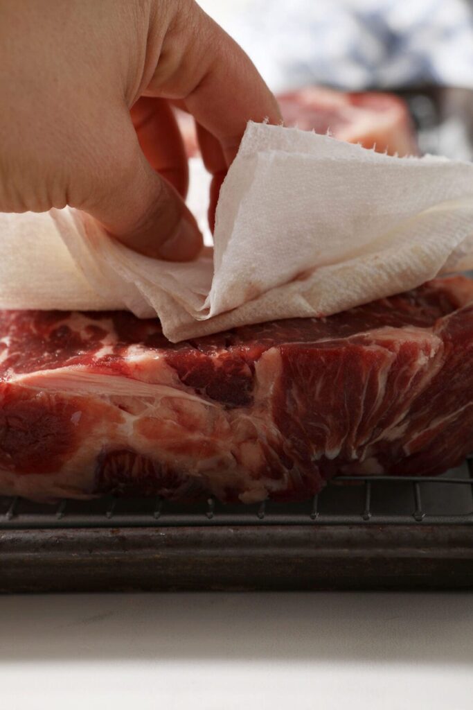 A hand blots a steak with a paper towel after seasoning