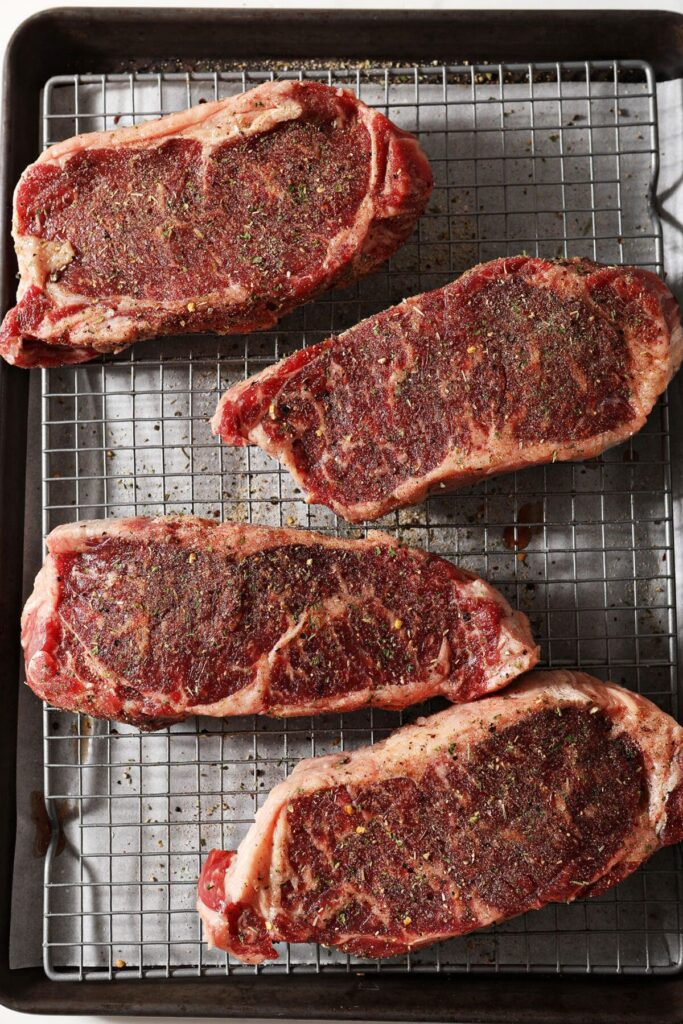 Four steaks on a wire cooling rack above a sheet pan seasoned with steak seasoning