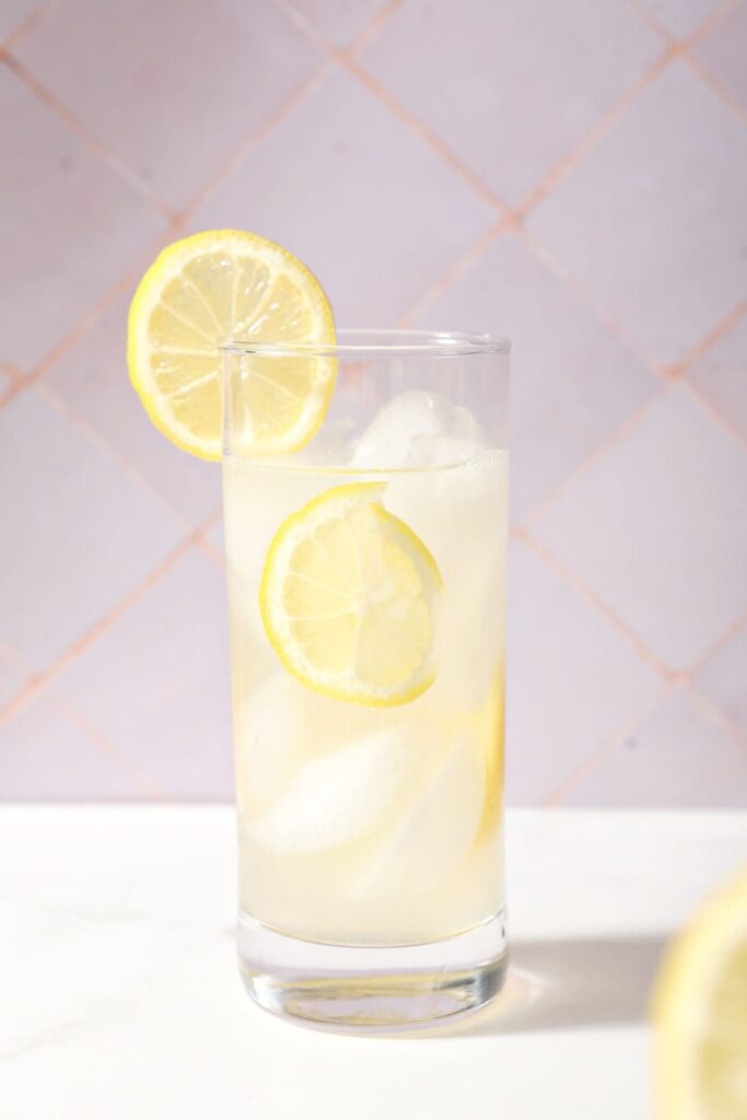 A glass of lavender lemonade garnished with a lemon wheel in front of a purple tile wall